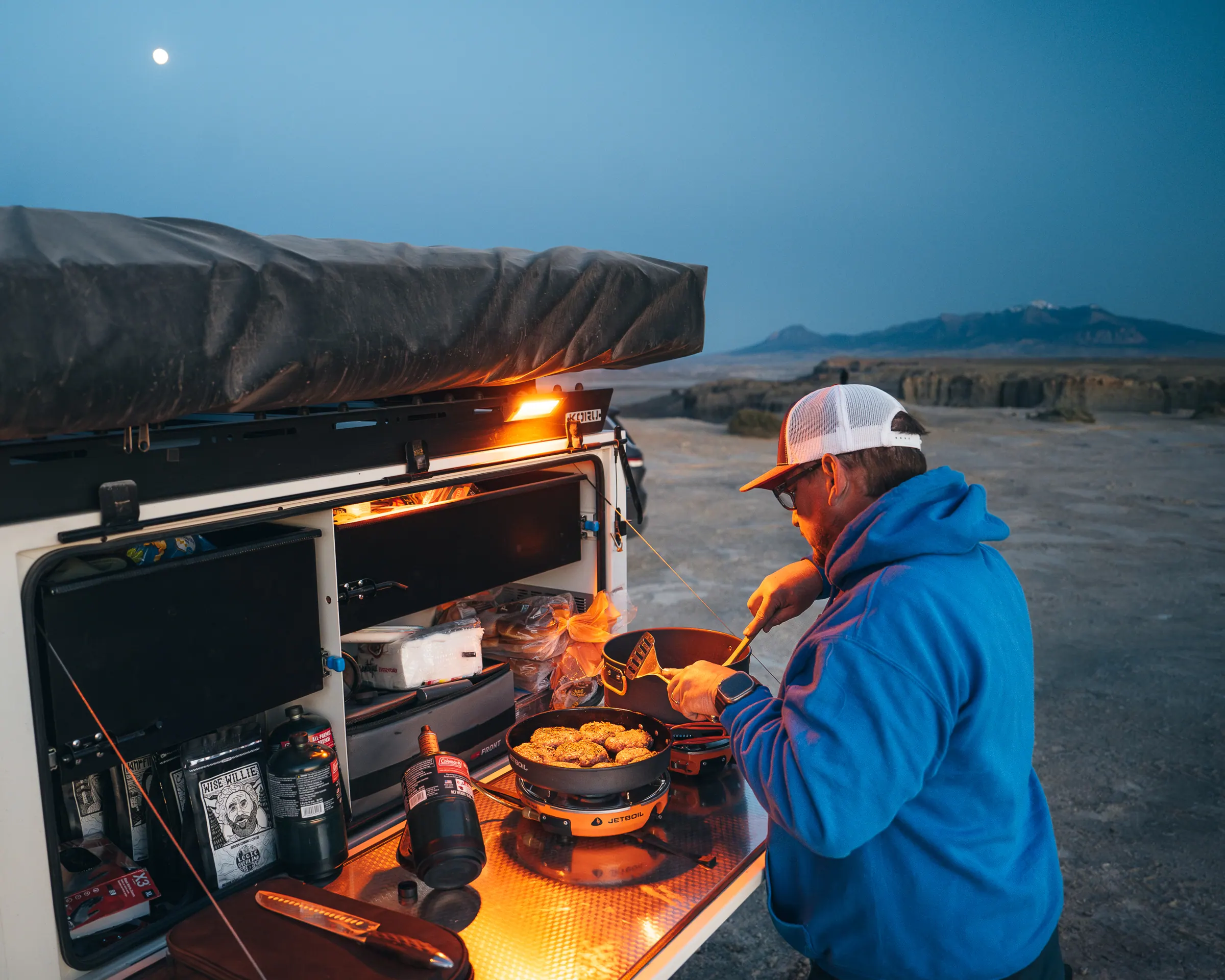 A camper cooking at the lit slide-out kitchen of an overland trailer at dusk in the desert