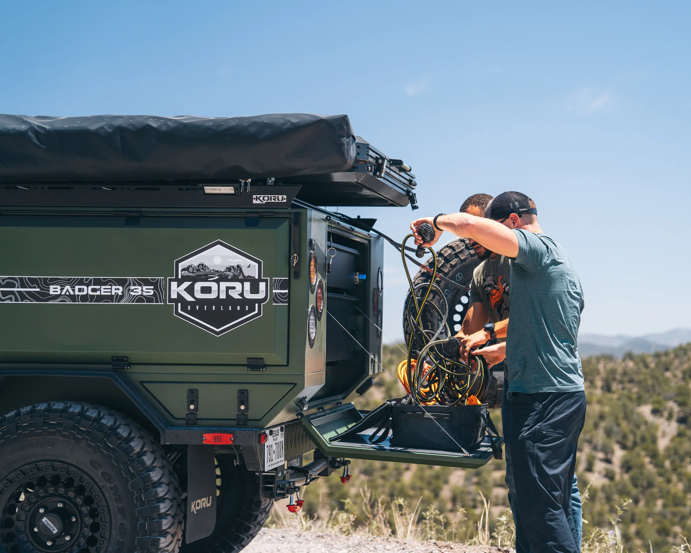 Two people working at the open side storage of a green Koru Badger 35 overland trailer on a mountain trail